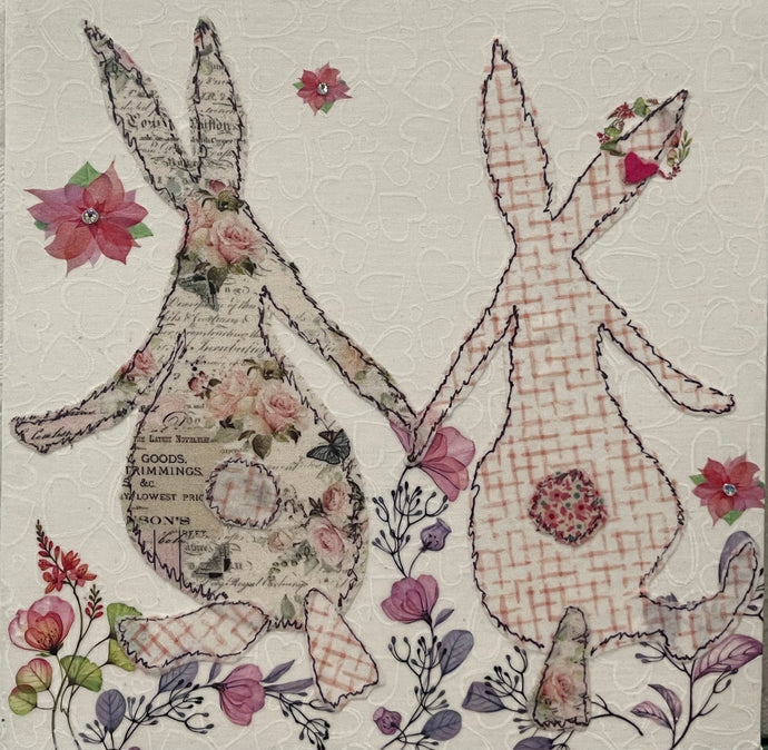 Whimsical bunny artwork made with layered fabric and mixed media, featuring paired figures in soft neutral and blush tones with textured stitched looking details running through the flowers
