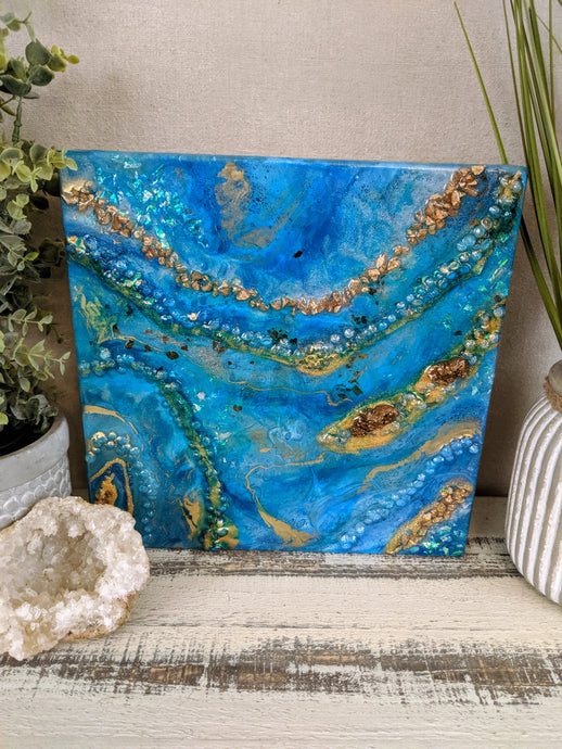 Abstract artwork made with resin and recycled glass on canvas, featuring deep blue and turquoise tones with gold accents and textured reflective surface